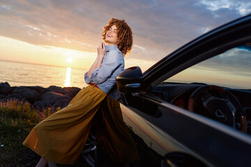 Fototapeta premium Cheerful woman enjoying outdoor drive beside a sleek car at sunset, leaning against the door. Lifestyle moment emphasizing fashion, confidence, and relaxed happiness.