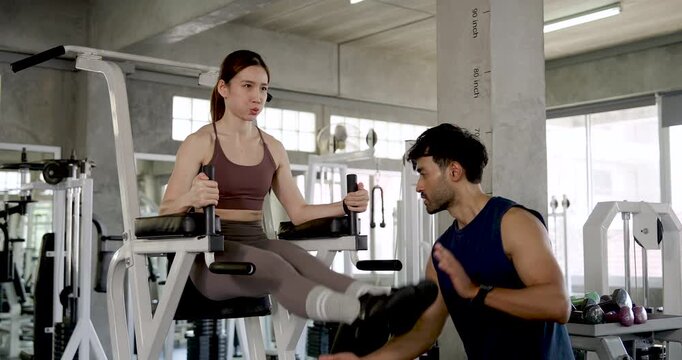 Young Asian couple exercising together in modern gym, woman performs knee raise on fitness machine while man guides movement, partner leg training with support, motivation, teamwork, wellness routine.