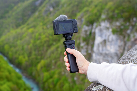 Close-up of a hand holding a compact action camera on a gimbal above a lush green gorge, outdoor content creator moment