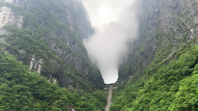 Tourists climbing steep staircase toward massive natural limestone arch in mountain. Tianmen mountain heaven gate cave with lush green forest. Scenic Asian landmark and outdoor travel adventure.