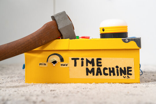 Yellow handmade Time Machine box with an axe wedged into its top on a white surface, humorous destruction concept, creative prop, quirky art.