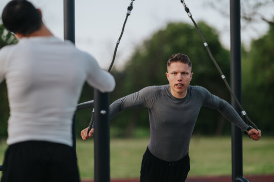 A young man performs a suspension cable chest fly exercise while training with a partner. They work out together on outdoor gym equipment in a park setting.