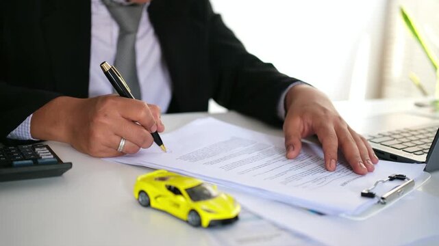 Man signing car insurance document or lease paper. Writing signature on contract or agreement. Buying or selling new or used vehicle. Car keys on table on red car background. Warranty or guarantee.
