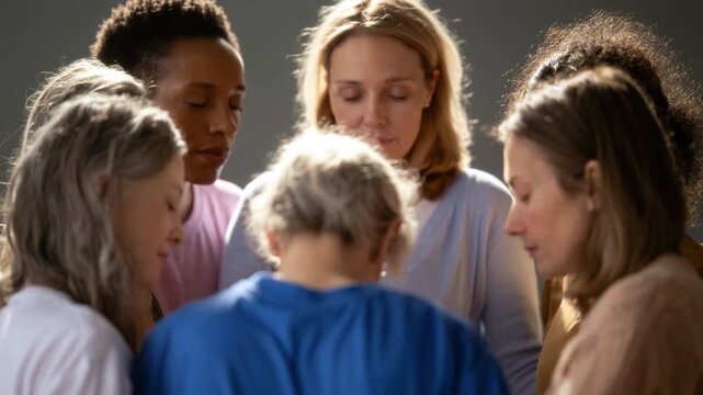 Group of diverse women standing closely together with eyes closed in a moment of support and solidarity indoors with soft lighting and muted background