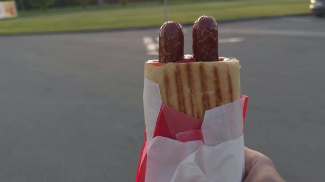 Hand holding grilled hot dog with two sausages, wrapped in paper and paper cone, served outdoors beside empty road in bright sunlight, close up food street snack with casual summer feel