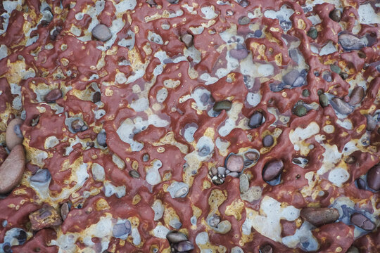 Abstract natural texture of red laterite rock with honeycomb weathering pattern and embedded multi-colored pebbles on the beach, geological background.