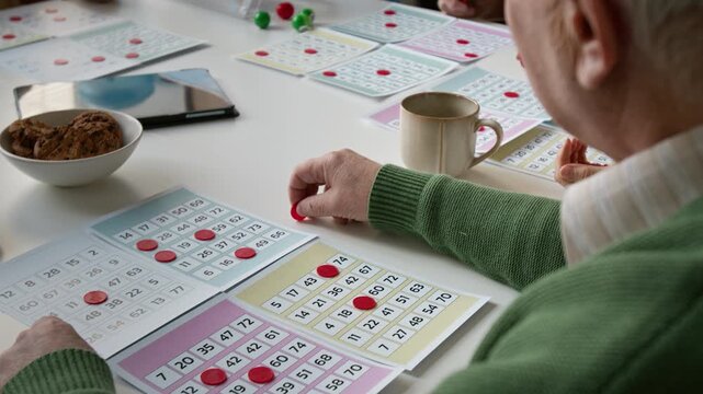 Over shoulder shot of senior man playing bingo in retirement home, placing red chips on card. Video for promoting elderly care services and cognitive health programs