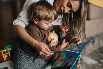 A mother reads a colorful board book to her toddler as they sit on the floor. The parent and child...