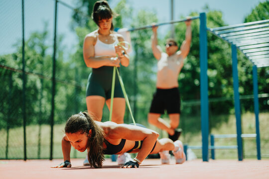 A fit woman performs push-ups with a resistance band while teammates work out in the background. Outdoor group training in a park features bodyweight and calisthenics exercises on a sunny day.