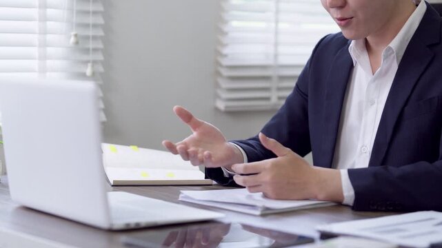 Asian businesswoman gesturing in video conference call at home office desk with laptop and documents