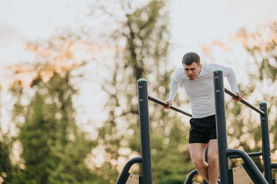 A man performs a dip exercise on parallel bars while training outdoors. He works on strength and balance during a park workout at sunset.
