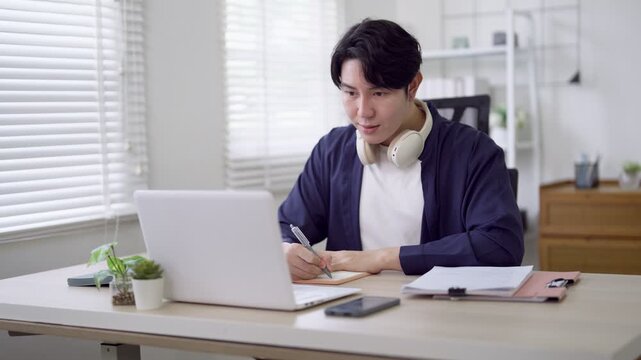 Young Asian freelance professional writing notes while working on laptop with headphones in bright home office