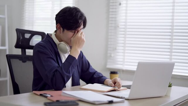 Young Asian freelancer feeling stressed while working remotely with laptop and headphones in bright modern home office