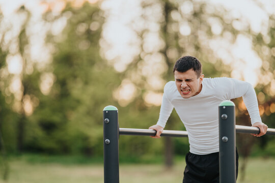 A man performs parallel bar dips outdoors, straining as he pushes his body upward. The athlete trains calisthenics in a park setting during an outdoor workout.