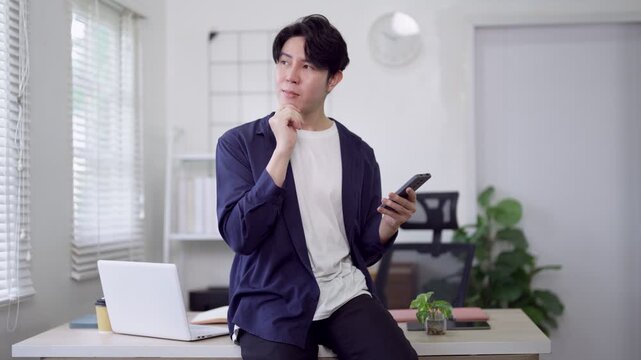 Young Asian freelance professional celebrating success message on smartphone while working from home office with laptop