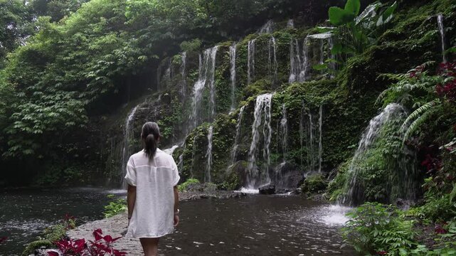 Woman in a white tunic admiring a lush tropical waterfall, enjoying the serene and wild nature
