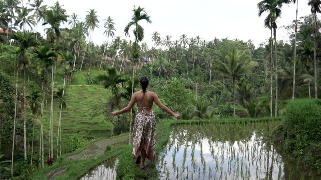 Following a woman in a backless dress walking beside lush green rice paddies in bali, indonesia