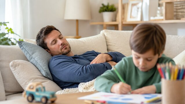 Man naps on a sofa while a young boy concentrates on coloring at a table in a cozy, softly lit living room