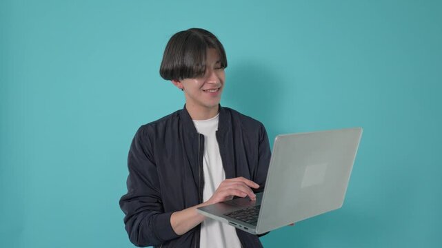 Cheerful young man standing and happily typing on his laptop computer against a solid turquoise background, showcasing modern communication, remote work, and positive digital interaction