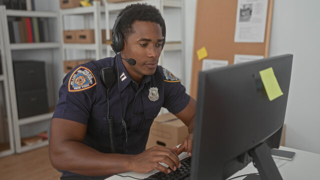 Man in police uniform wearing a headset typing on a keyboard at a building desk with monitor, badge, and cardboard boxes visible; focused duty.