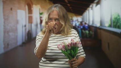 Fototapeta premium Senior woman points finger at camera while holding pink tulips and wearing striped shirt in a building corridor; defiance resolve.