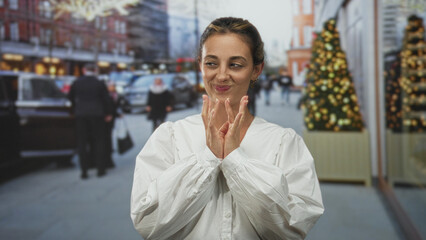Fototapeta premium Woman with hands clasped at chest wearing white blouse, smiling and looking up on street beside decorated storefront and christmas tree; wishful hope holiday shopping.