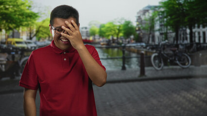 Young hispanic man laughing with hand over face on canal street in amsterdam wearing red polo and glasses; joyful travel.