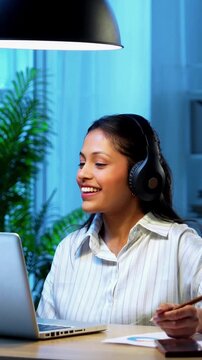 Indian businesswoman smiling attending video call on laptop using headphone at desk in modern cozy office, Asian young corporate executive expressing positivity while engaged in virtual meeting