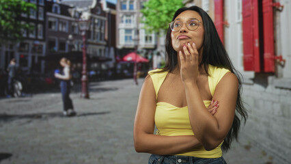 Fototapeta premium Woman wearing glasses with hand on chin and arms crossed, standing on a cobblestone street near red shutters; thoughtful contemplation.