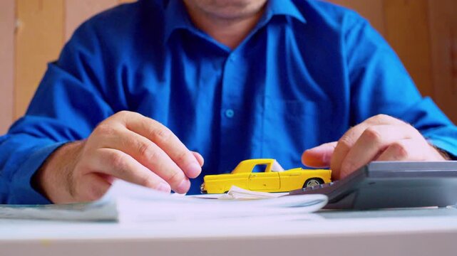 Car insurance agent playing with small yellow toy car at desk close up. Focus on hands and vehicle, shallow DOF, natural light. Bored businessman playing with miniature auto, procrastinating at work.