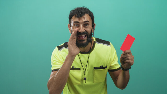 Man referee in neon shirt holds red card and whistle, hand cupped to mouth as if shouting, headset mic and beard visible in studio setting; discipline stern.