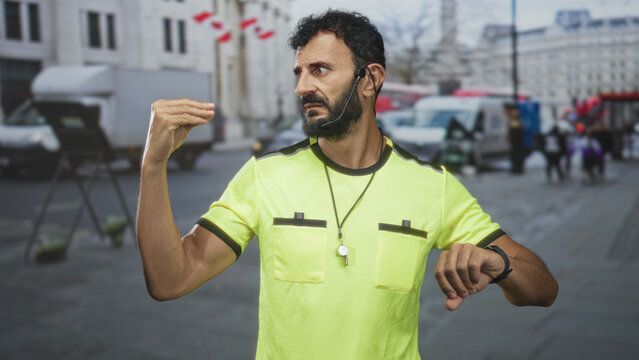 Man referee in yellow shirt wearing headset and whistle lanyard checks wristwatch and holds whistle amid parked vans and pedestrians on a busy city street; impatience.
