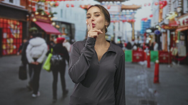 Woman finger to lips for silence in a crowded chinatown street, standing center frame with visible neck and casual zip top; mystery secrecy.
