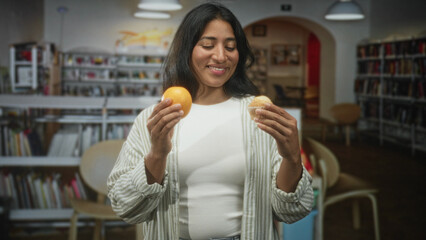 Young hispanic woman holding an orange and a muffin in her hands, smiling and comparing the snacks while standing among bookshelves in a public library building  choice contemplation. © Krakenimages.com
