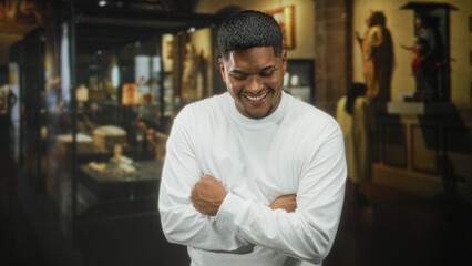 Fototapeta premium Hispanic man with beard crossing arms and smiling, hands visible in a museum building near glass display cases wearing white shirt; happiness warmth.