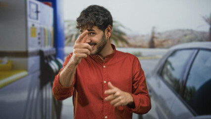 Smiling young hispanic man in red shirt points finger to camera near gas pump and car on street  playful confidence. © Krakenimages.com