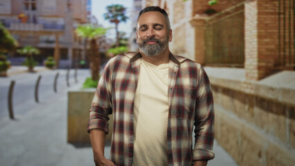 Obraz premium Middle age man with grey beard smiling with eyes half closed, standing on a sunny street plaza with hands relaxed at sides; contentment confidence.