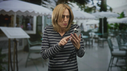 Fototapeta premium Woman tapping smartphone with visible hands and furrowed expression at a street cafe terrace, holding phone while scrolling through messages; everyday tech frustration.