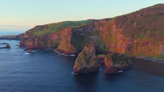 Aerial view of the rugged coastal cliffs and rock formations at Miradouro dos Caimbros, featuring lush green hills and golden hour light over the Atlantic Ocean in Cedros, Acores, Portugal.