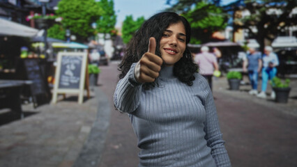 Fototapeta premium Young woman smiling and giving thumbs up toward camera on a busy pedestrian street by outdoor cafe and chalkboard sign; optimism confidence.