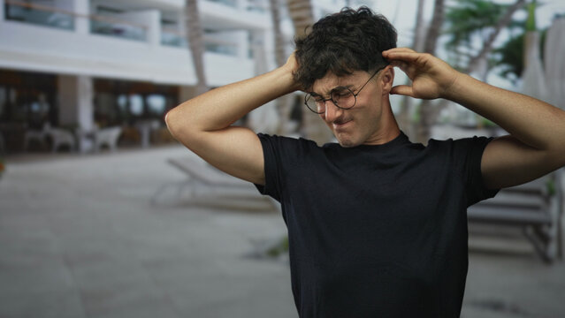 Man with hands on head and bare forearms visible wearing glasses and black tshirt at building poolside; frustration reaction.