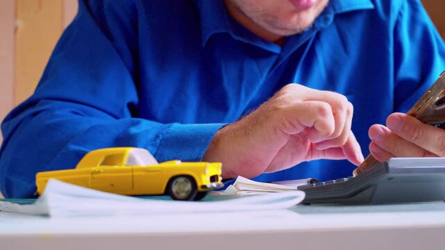 Car insurance agent calculating cost on smartphone at desk close up. Focus on hands, yellow toy car on table. Shallow depth of field, natural light. Businessman estimating price of vehicle policy.