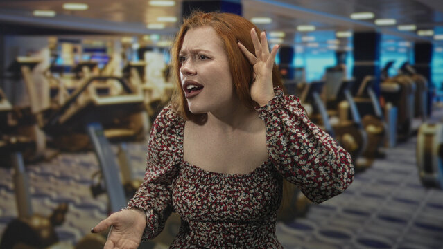 Redhead woman cups ear and points finger toward camera amid gym cardio machines in building, leaning forward with puzzled frown and raised hand; frustration confrontation.