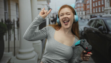 Redhead woman wearing cat headphones holds a gamepad while raising her arm and making a rock sign on a street  joy celebration. © Krakenimages.com