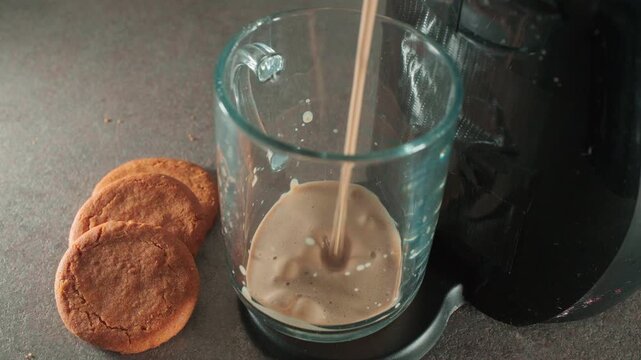 Empty glass sits beside cookies on a table. Liquid pours into glass, creating frothy bubbles. Dark machine beside glass suggests coffee preparation. Warm, comforting drink with snacks nearby.
