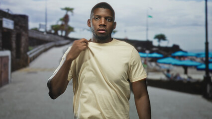 Fototapeta premium Man pulls shirt at neck, exposing part of chest and wearing a tense expression on a beachside street lined with umbrellas and palm trees; discomfort.