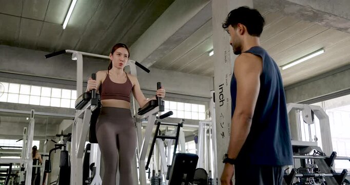 Young Asian couple training together in modern gym, woman lifts knees on vertical raise station while man watches and encourages, partner workout with leg strength, support, wellness motivation.