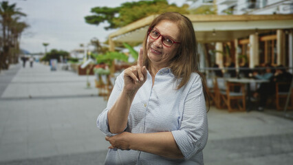 Fototapeta premium Woman points finger up at chest level on a street beside an outdoor cafe, wearing red glasses and a striped shirt; gentle advice warmth.