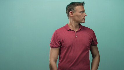 Fototapeta premium Man in red polo scratches neck with hand behind neck in studio teal backdrop, visible forearm and pensive expression; thoughtful uncertainty.