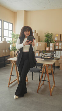 Woman taps tablet screen with hands amid stacked cardboard boxes in an office building while checking parcels and inventory; determination.